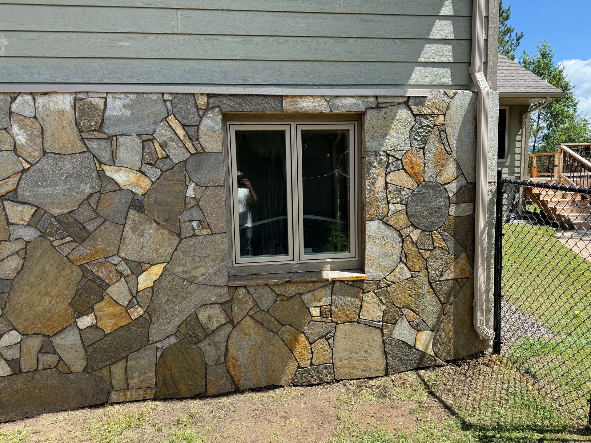 Stone exterior wall with window, next to a black fence and greenery.