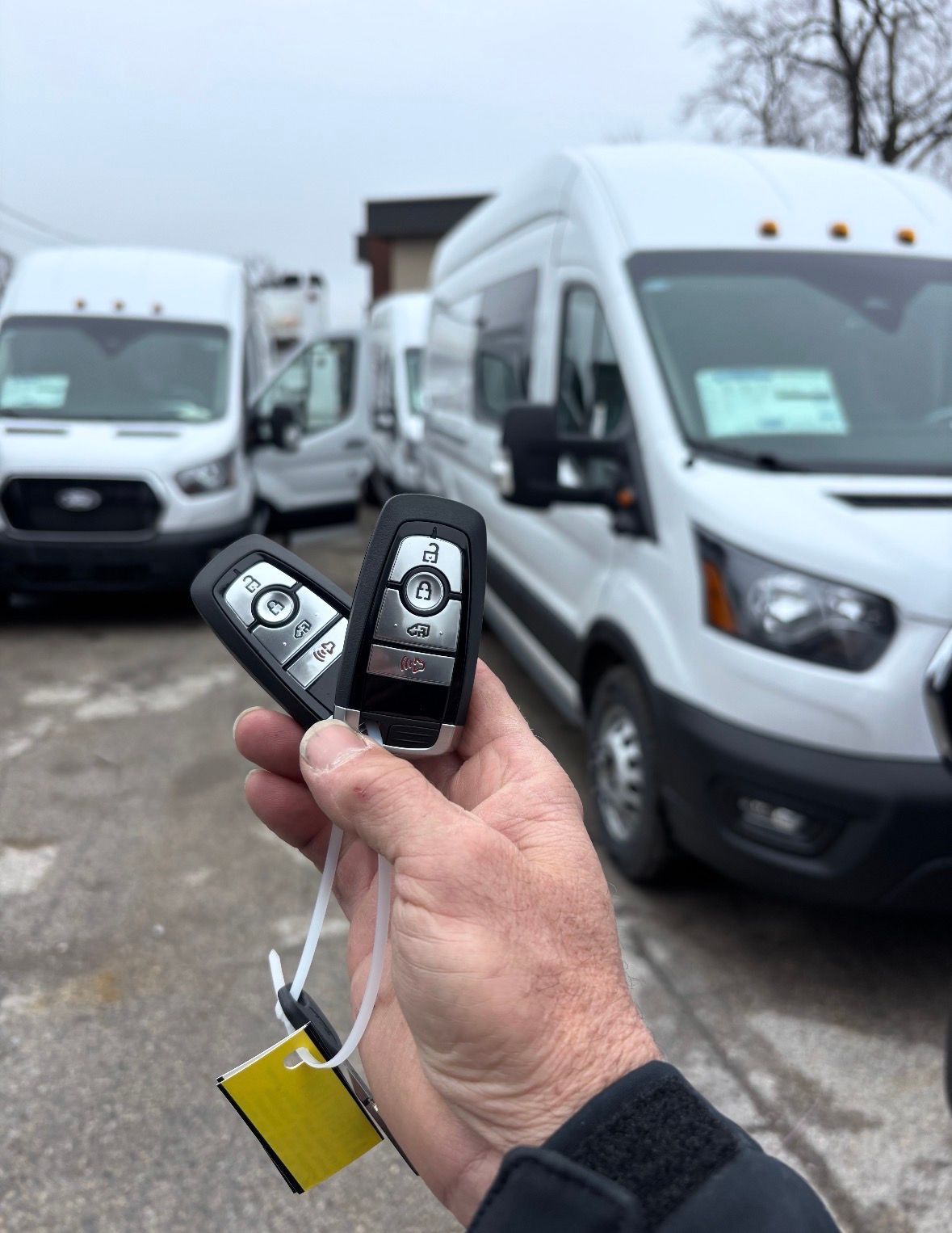 Hand holding two black key fobs in front of parked white vans on an overcast lot