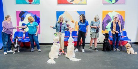 People with therapy dogs on leashes at an indoor event with colorful painted dog portraits.
