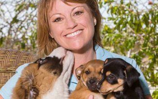 Smiling woman holding three puppies outdoors, with one puppy licking her cheek.
