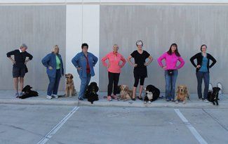 People and dogs lined up outdoors, standing with hands on hips in front of a wall.