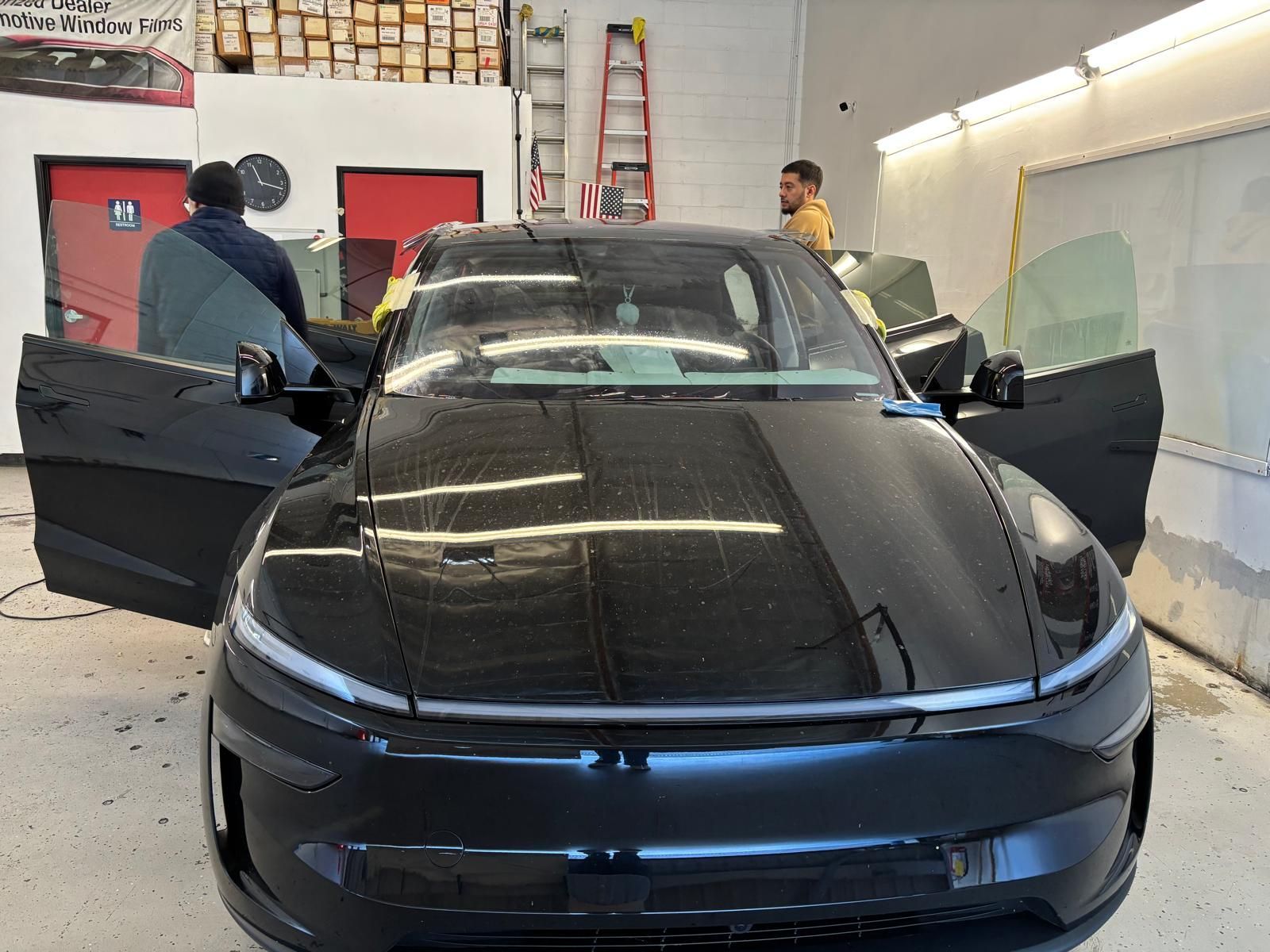 Two people installing tinted window film on a black car in a shop.