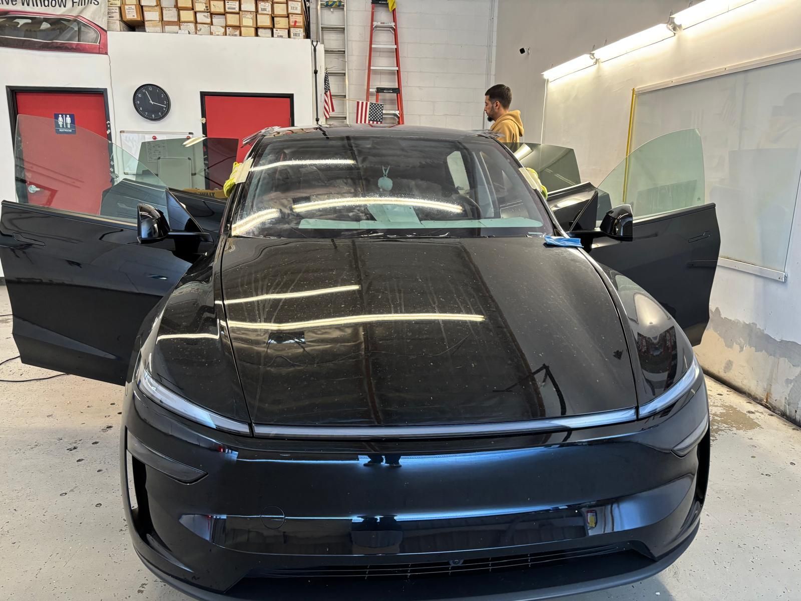 Black car with open doors inside a shop; a person is working on the windows.