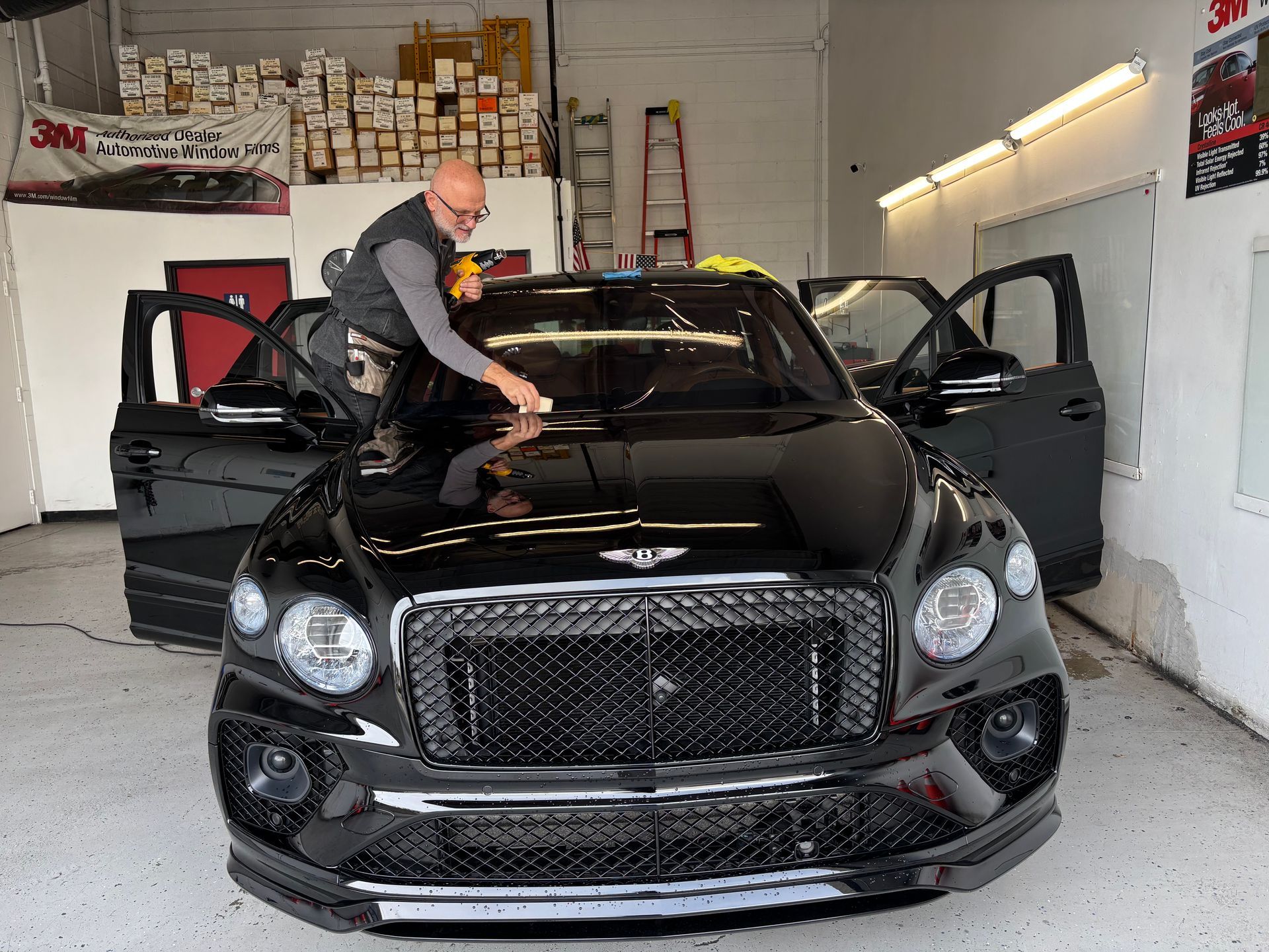Man applying tint to black Bentley SUV windshield in a shop.
