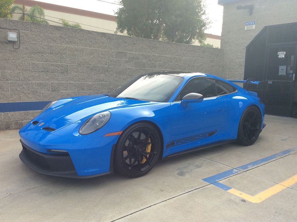 Blue Porsche sports car parked next to a brick building. Black wheels, yellow brake calipers, and a black roof.