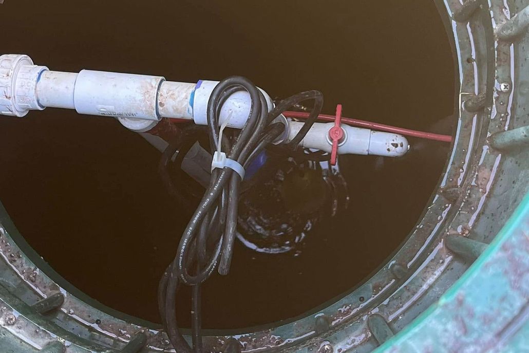 Inside a dark tank, white PVC pipe, black cables, and a float switch are visible. Green rim surrounds the opening.