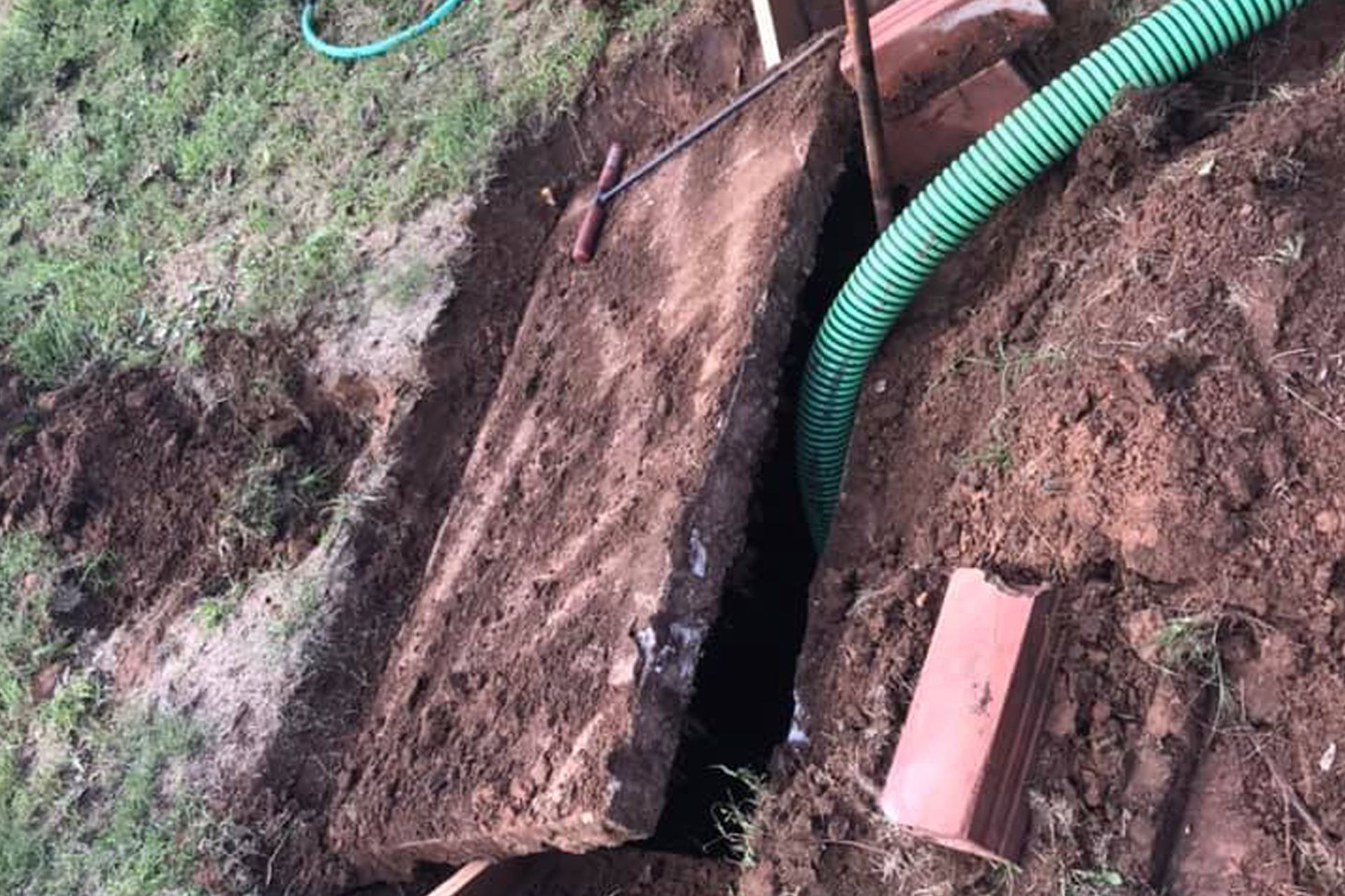 Excavated area with large stone slab and a green hose, alongside red bricks on the ground.