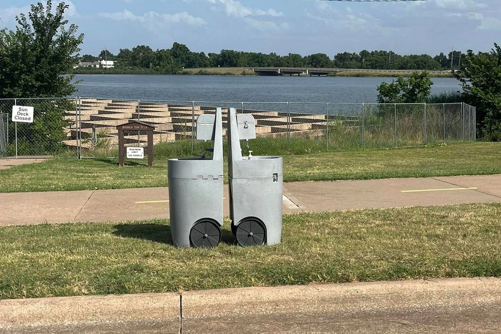 Gray robotic trash can on wheels outdoors near a lake and fence.