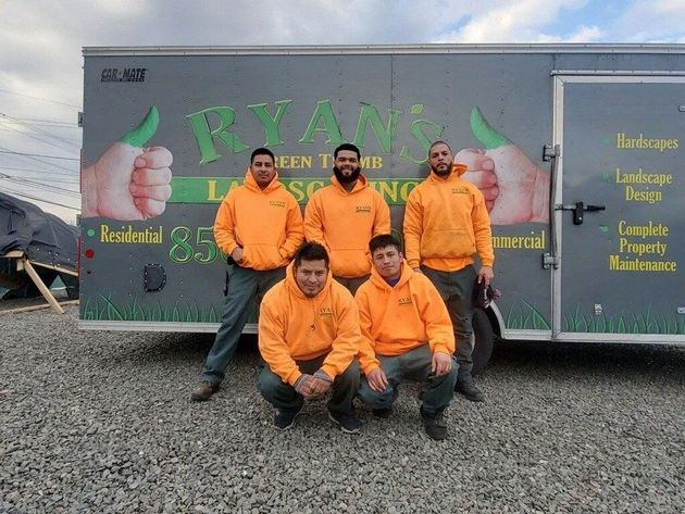 Five workers in bright orange hoodies stand and crouch in front of a gray Ryan’s Green Thumb landscaping trailer.