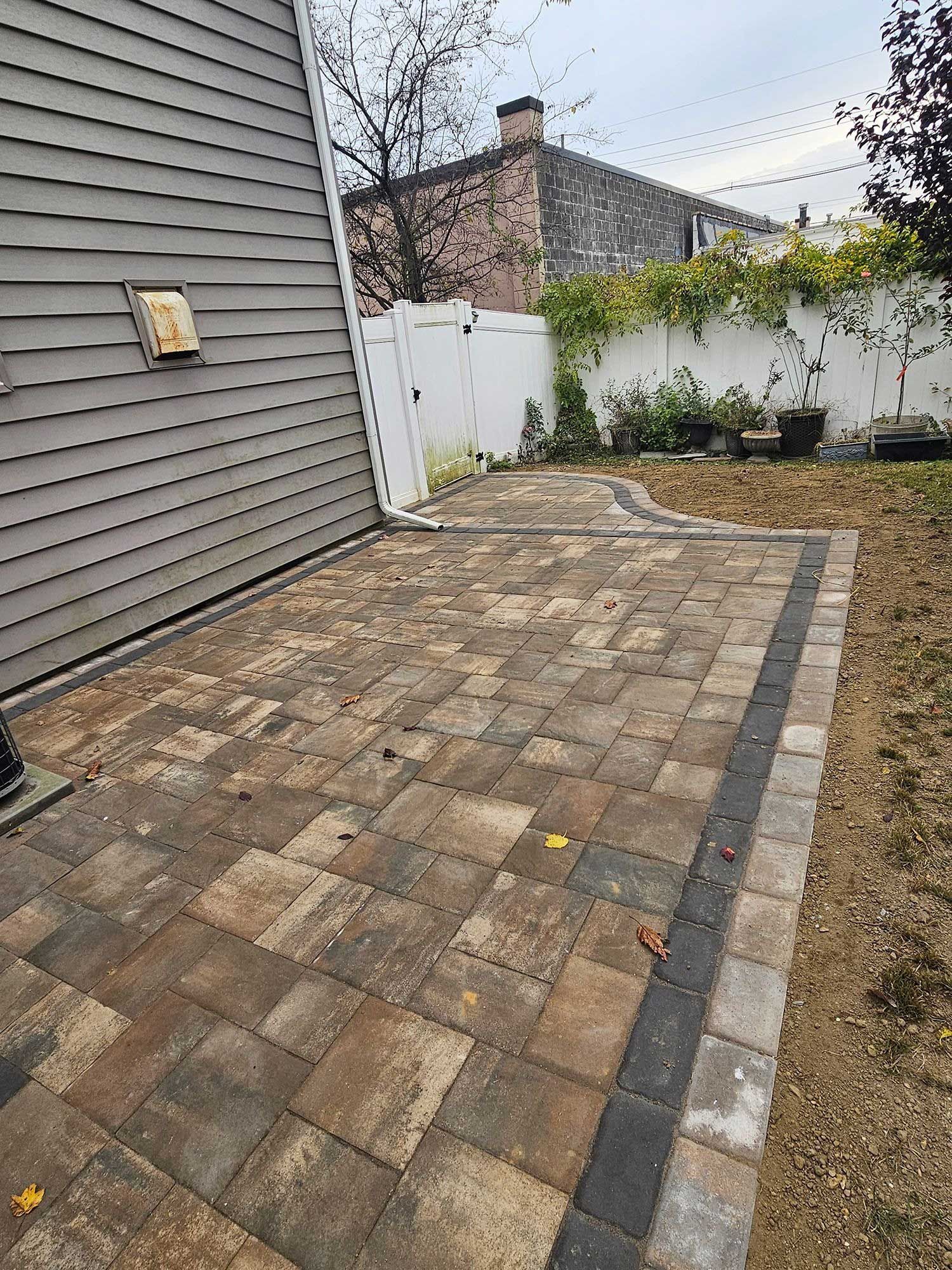 A multi-toned stone patio with a dark grey border running alongside a house with light grey vinyl siding.