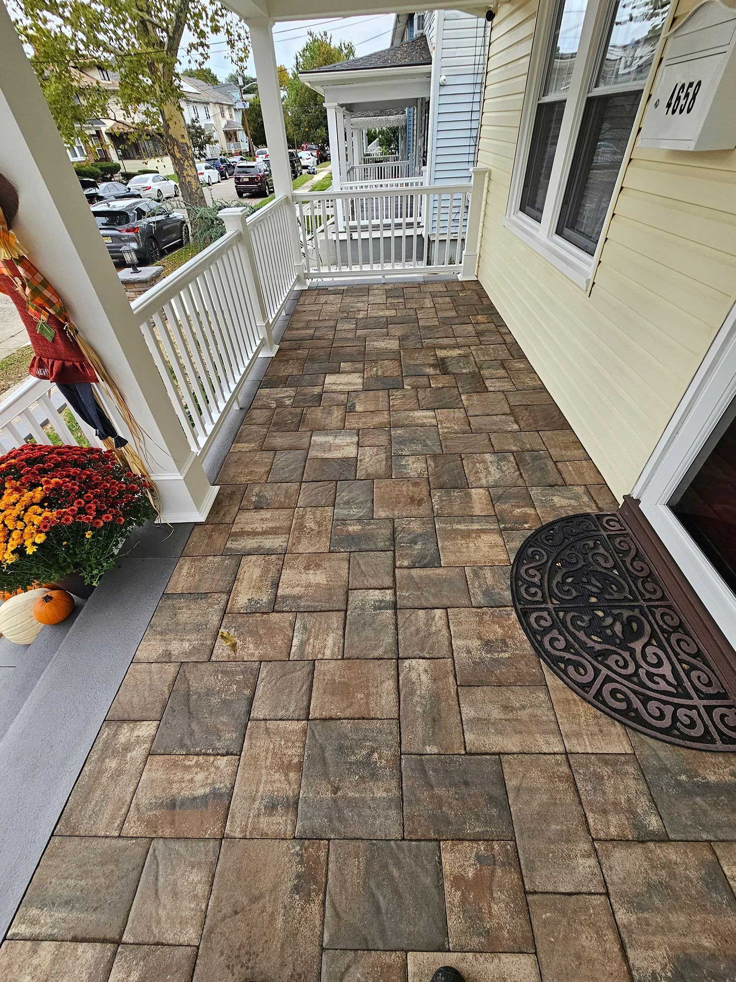 A porch with patterned, earth-toned stone pavers, white railings, and a decorative mat in front of a house door.