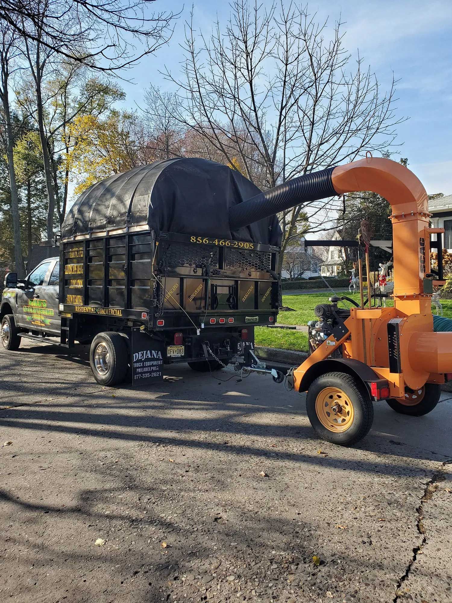 A black truck pulling an orange leaf vacuum trailer on an outdoor residential street.