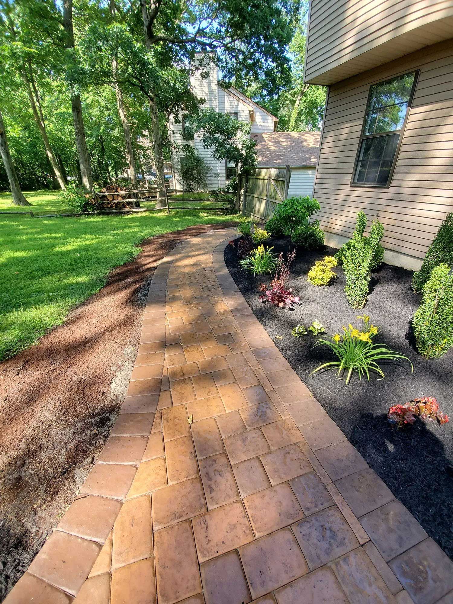 A curved brick paver walkway leads along the side of a house with a grass lawn and landscaped garden beds.