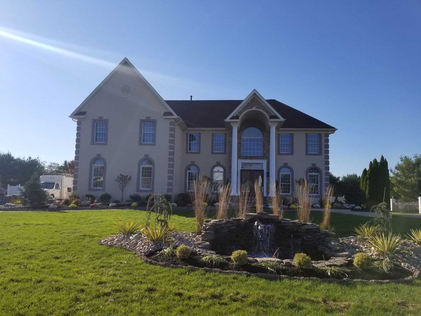 A large, light-colored two-story house with a lawn, landscaping, and a small stone waterfall feature in the foreground.