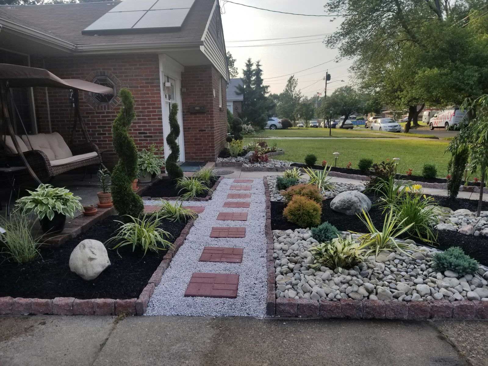 A brick-paved path with stone borders leads to a house entrance, flanked by garden beds with shrubs, rocks, and mulch.