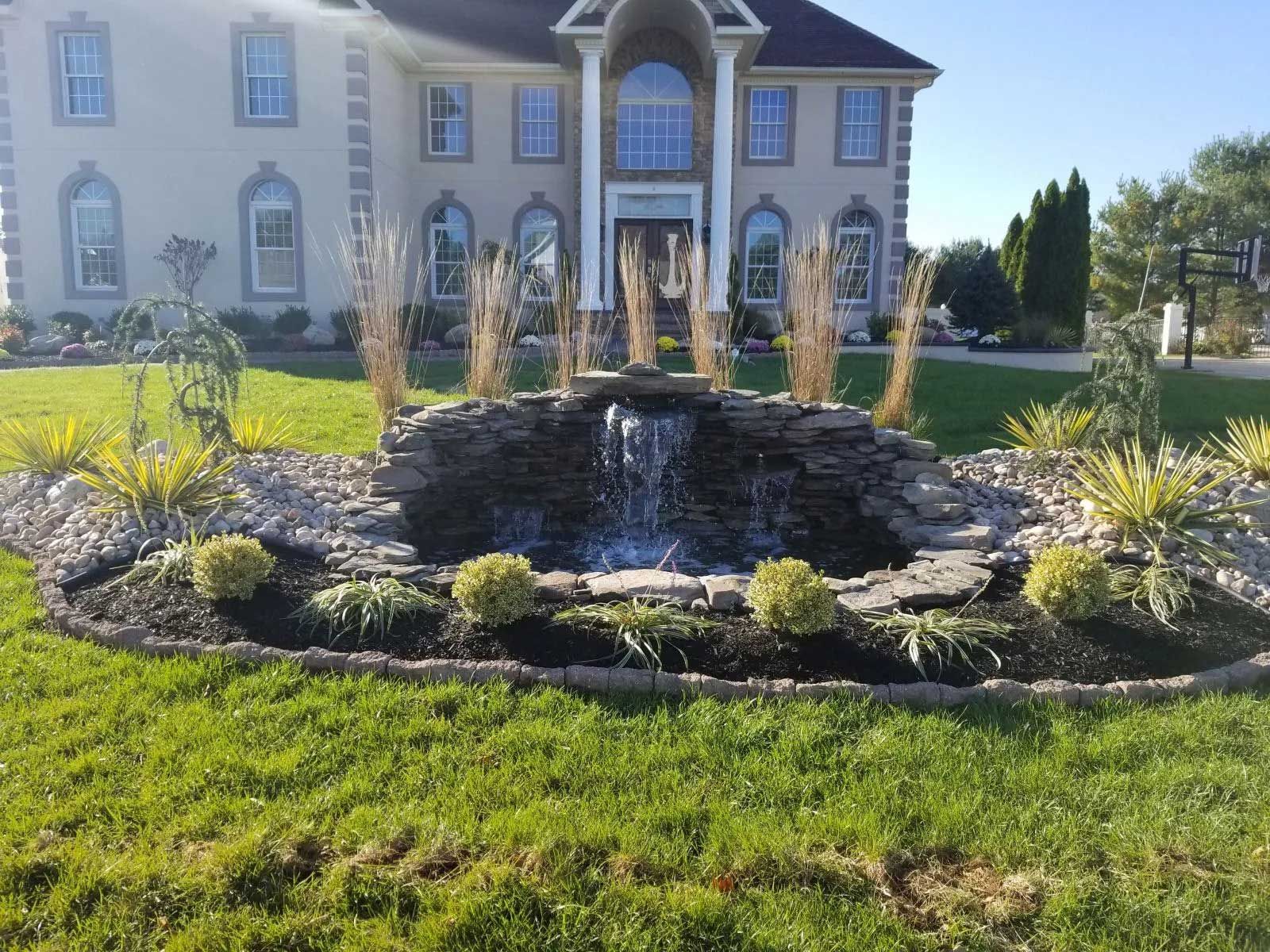 A landscaped rock waterfall feature sits in a front yard in front of a two-story beige house on a sunny day.