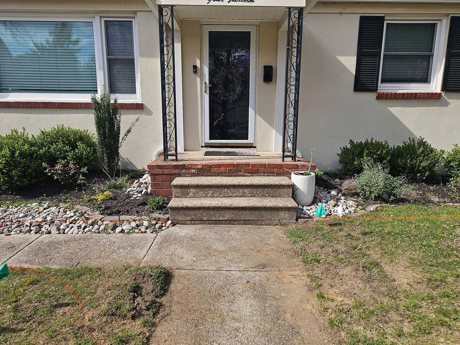 Front entrance of a light-colored house with a glass storm door, concrete steps, and landscaping.