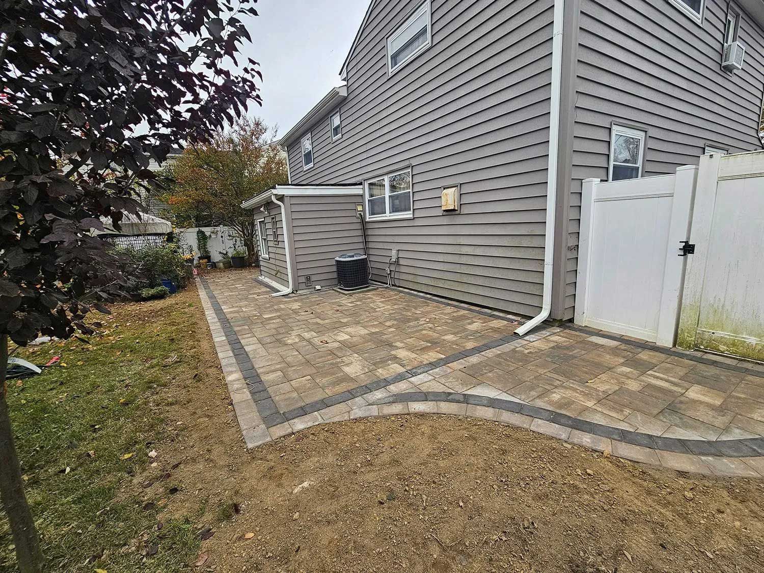 A gray house with a new paved patio featuring tan and dark gray stones next to a white fence in a backyard.