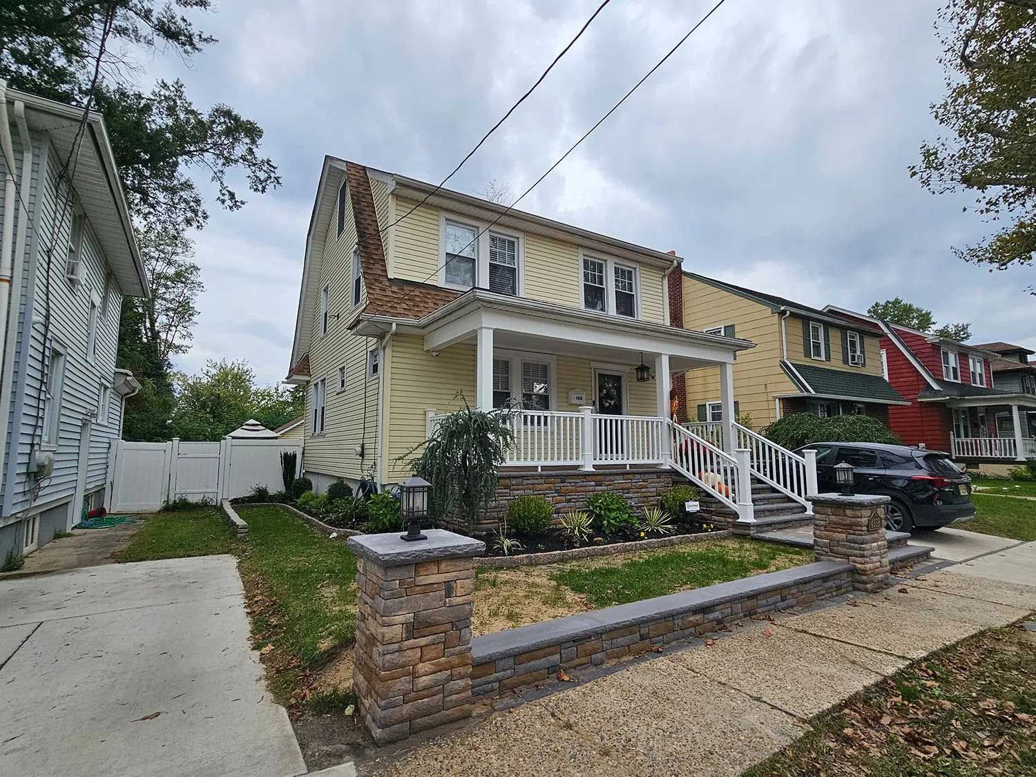 A two-story, yellow-sided house with a front porch, a stone retaining wall, and a concrete driveway on an overcast day.
