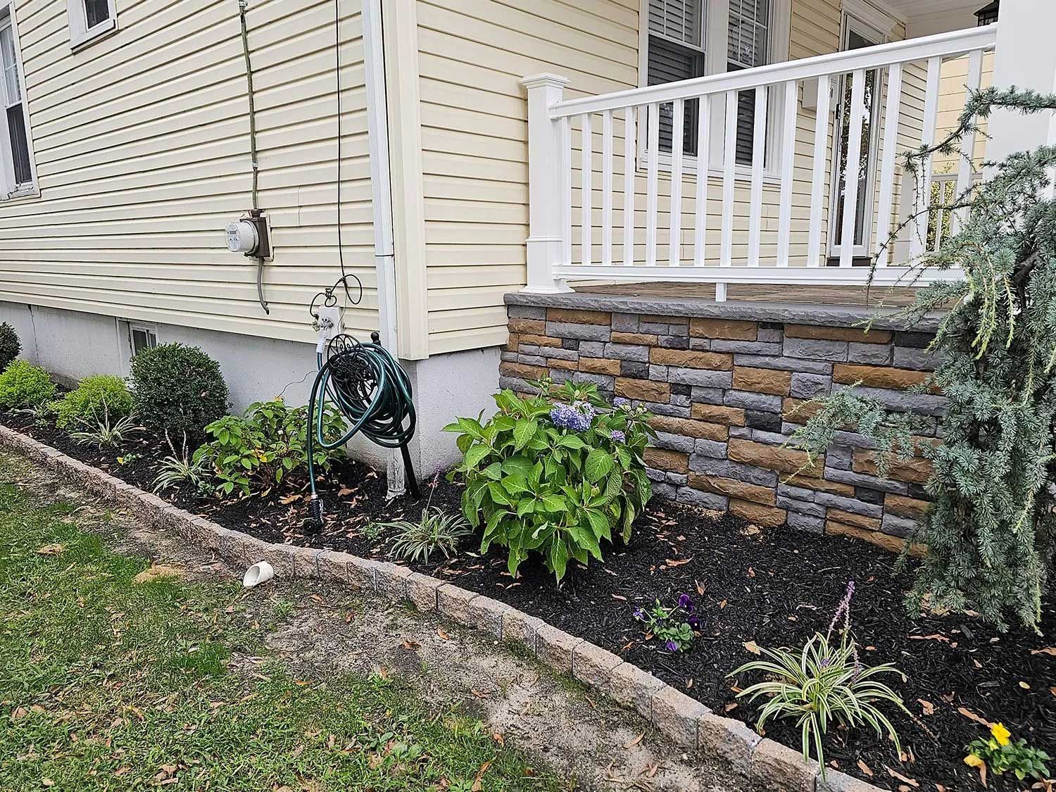 A garden bed with mulch and assorted green shrubs against a house with yellow siding and a stone-covered porch base.