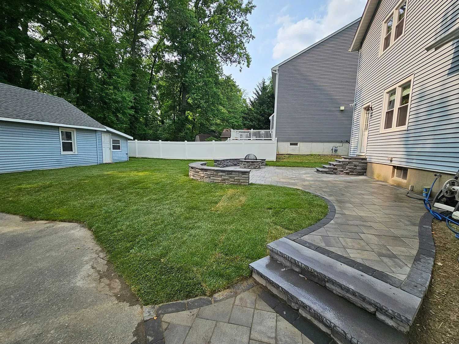 A backyard features a stone patio with steps, a stone fire pit, a green lawn, and white fencing, next to two houses.