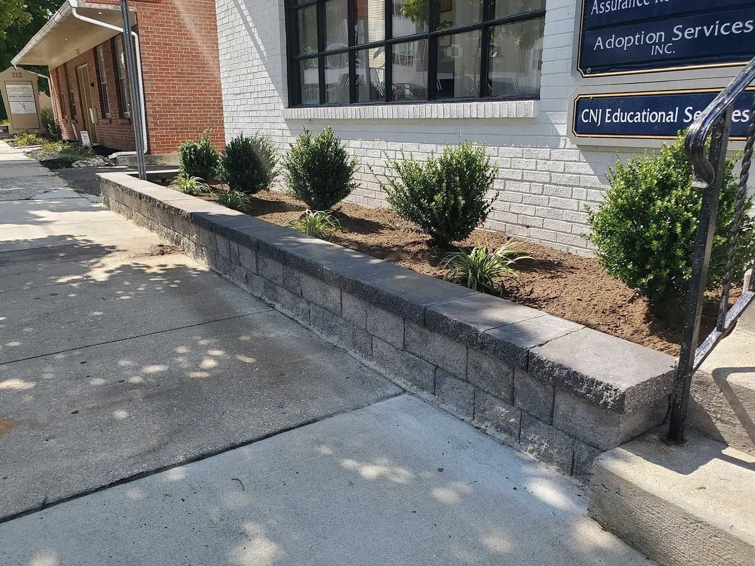 A low, gray stone retaining wall holds a garden bed with several small evergreen shrubs in front of a brick building.