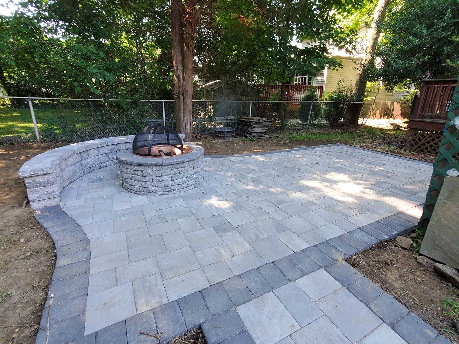 A patio with a stone fire pit and curved seating wall, surrounded by light gray pavers with a dark gray stone border.