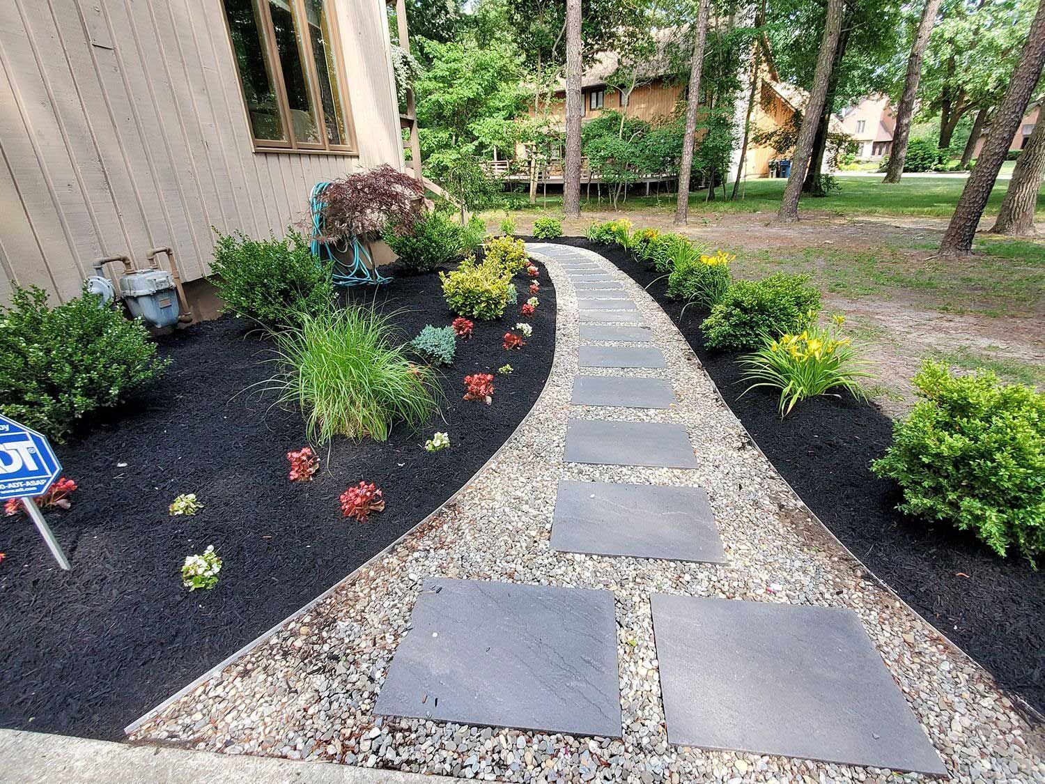 A stone pathway leads past landscaped garden beds filled with dark mulch and green shrubs against a beige house exterior.