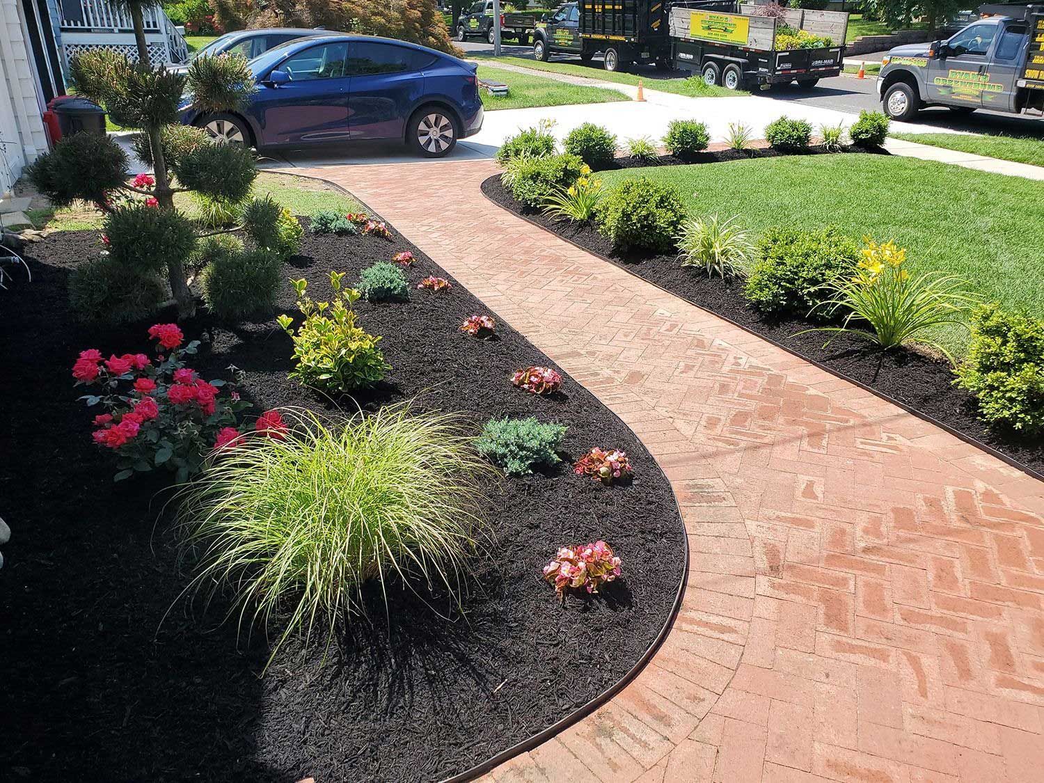 A winding brick pathway bordered by black mulch, green shrubs, and red flowers leads toward a blue car in a residential yard.
