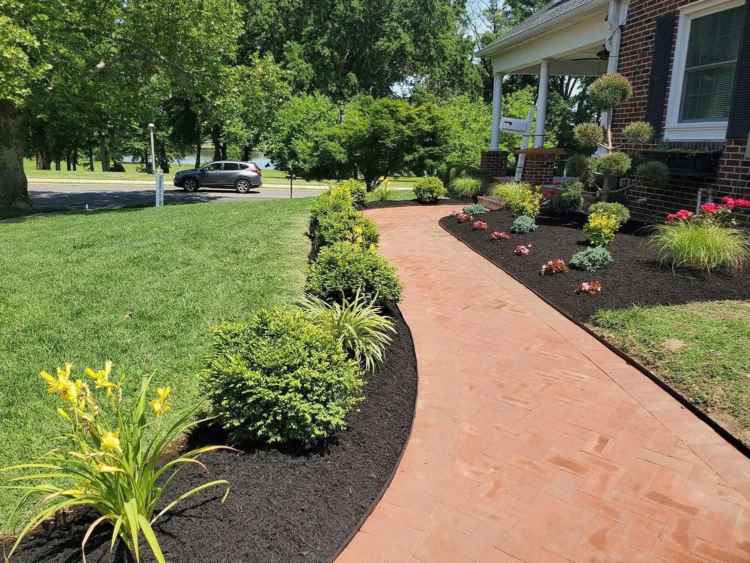 A curved, reddish walkway leads to a house entrance, lined with dark mulch, green shrubs, and yellow flowers in a lawn.