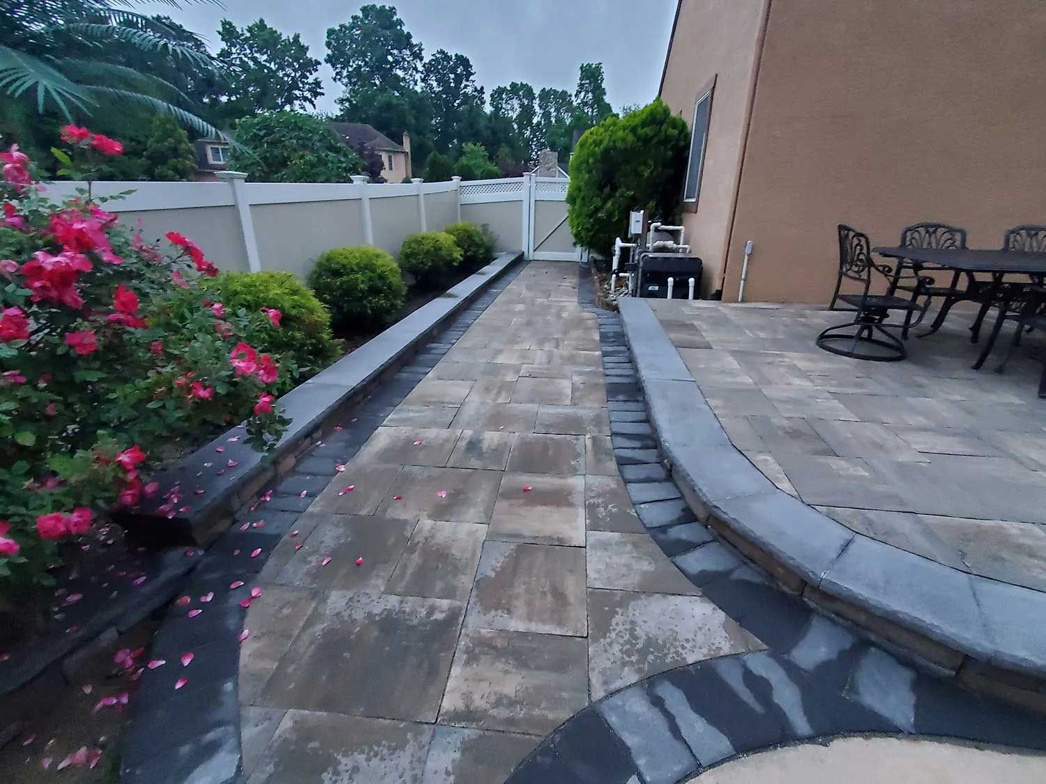 A stone walkway leads to a patio with outdoor furniture, flanked by a rose bush, trimmed shrubs, and a white fence.