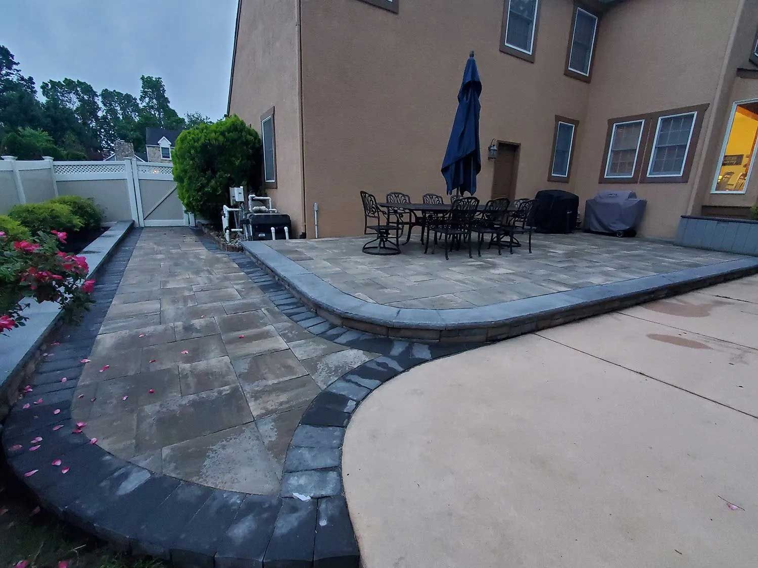 A backyard patio with light-colored pavers, a dark-edged border, outdoor furniture, and a closed umbrella by a tan house.