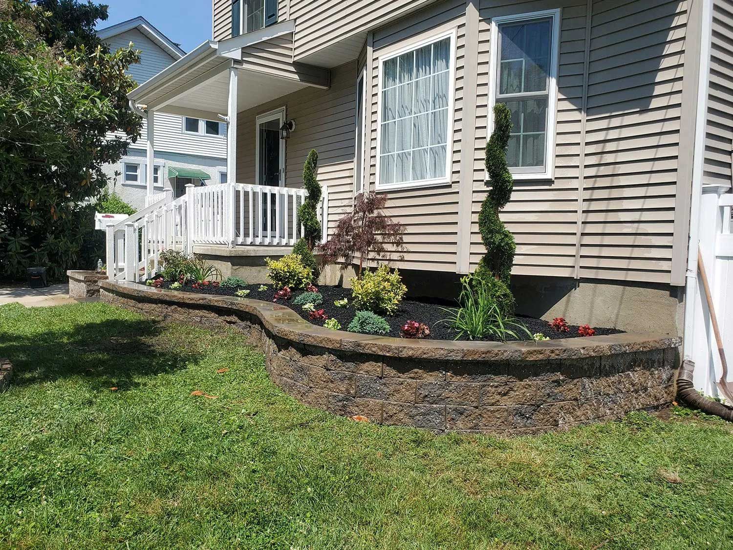 A house exterior featuring a curved stone retaining wall with mulch and various shrubs in front of light beige siding.