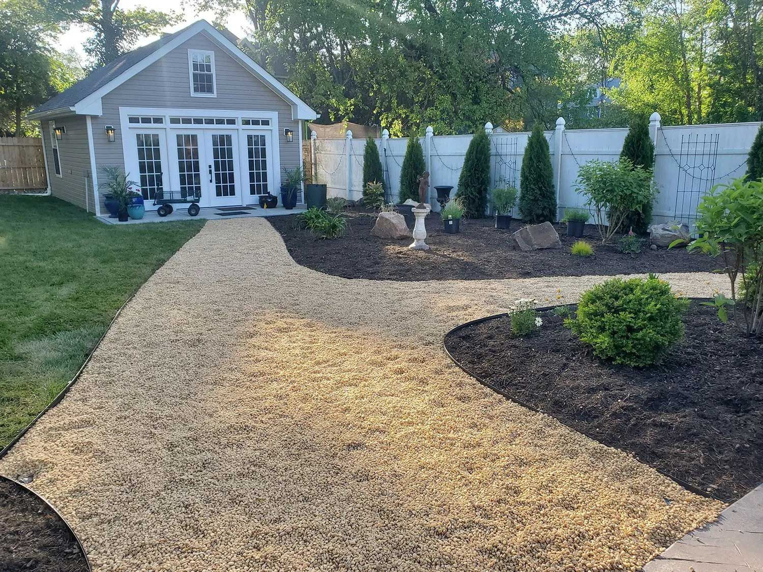 A light gray garden shed with French doors sits at the end of a winding tan woodchip path in a landscaped yard.
