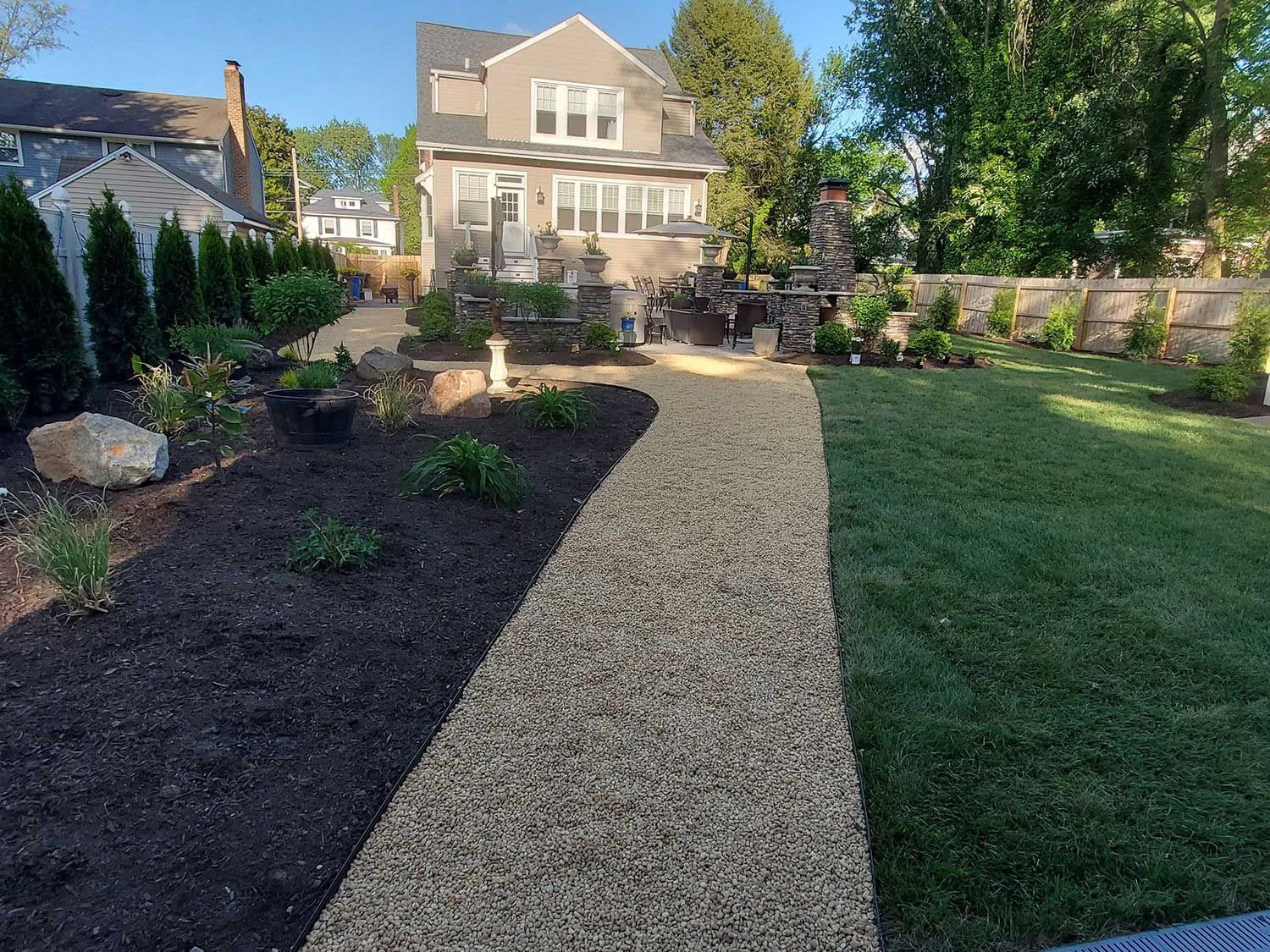 A gravel path leads to a house with a landscaped yard featuring dark mulch, new plants, and a green grass lawn.