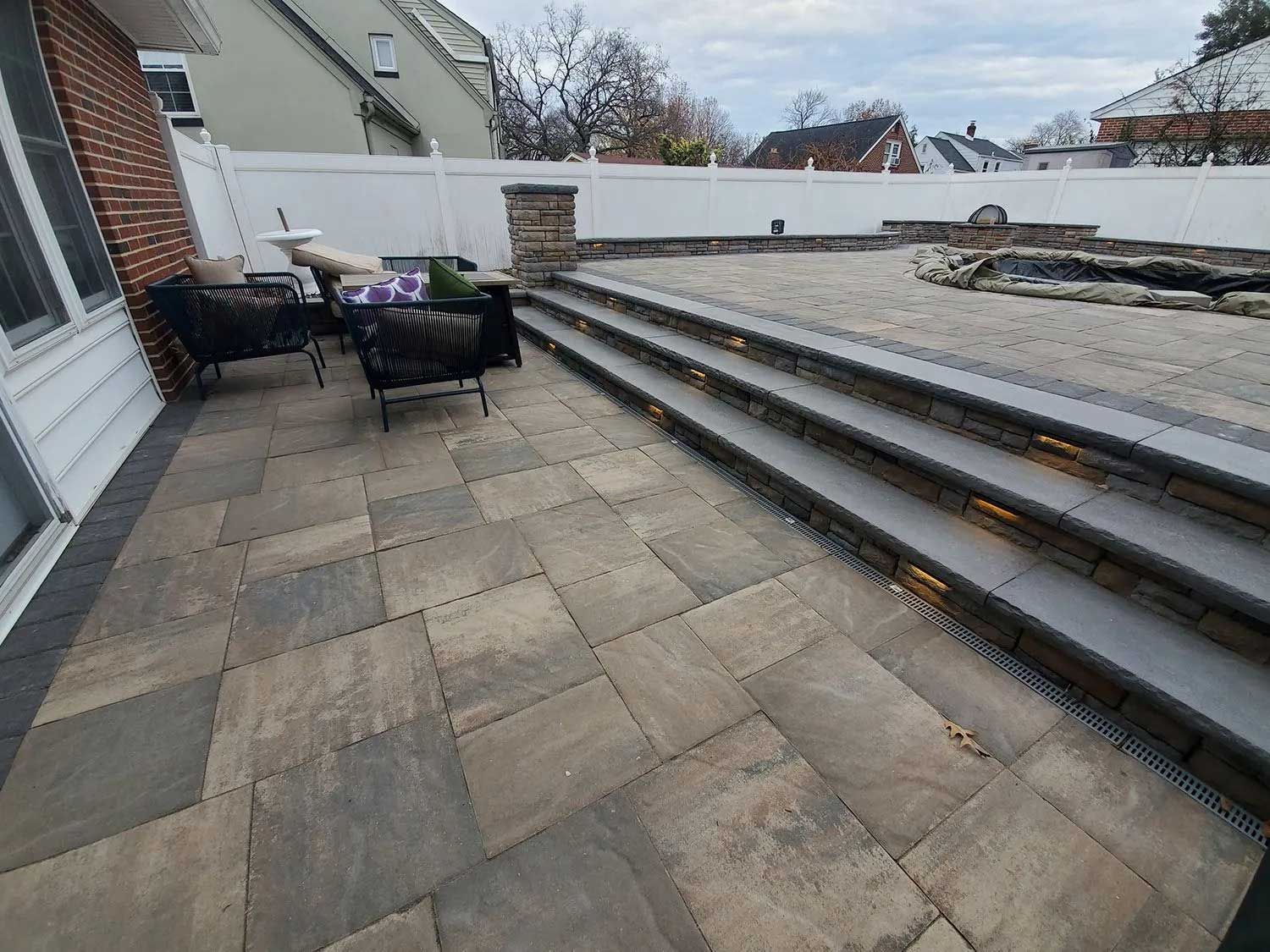 A paved patio with outdoor furniture leading up stone steps to a raised backyard area featuring a fire pit and white fence.