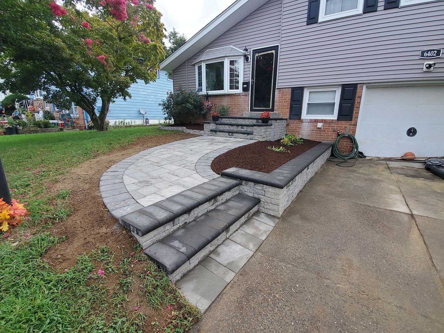 A newly installed stone patio and steps with a small raised garden bed lead to the entrance of a suburban home.