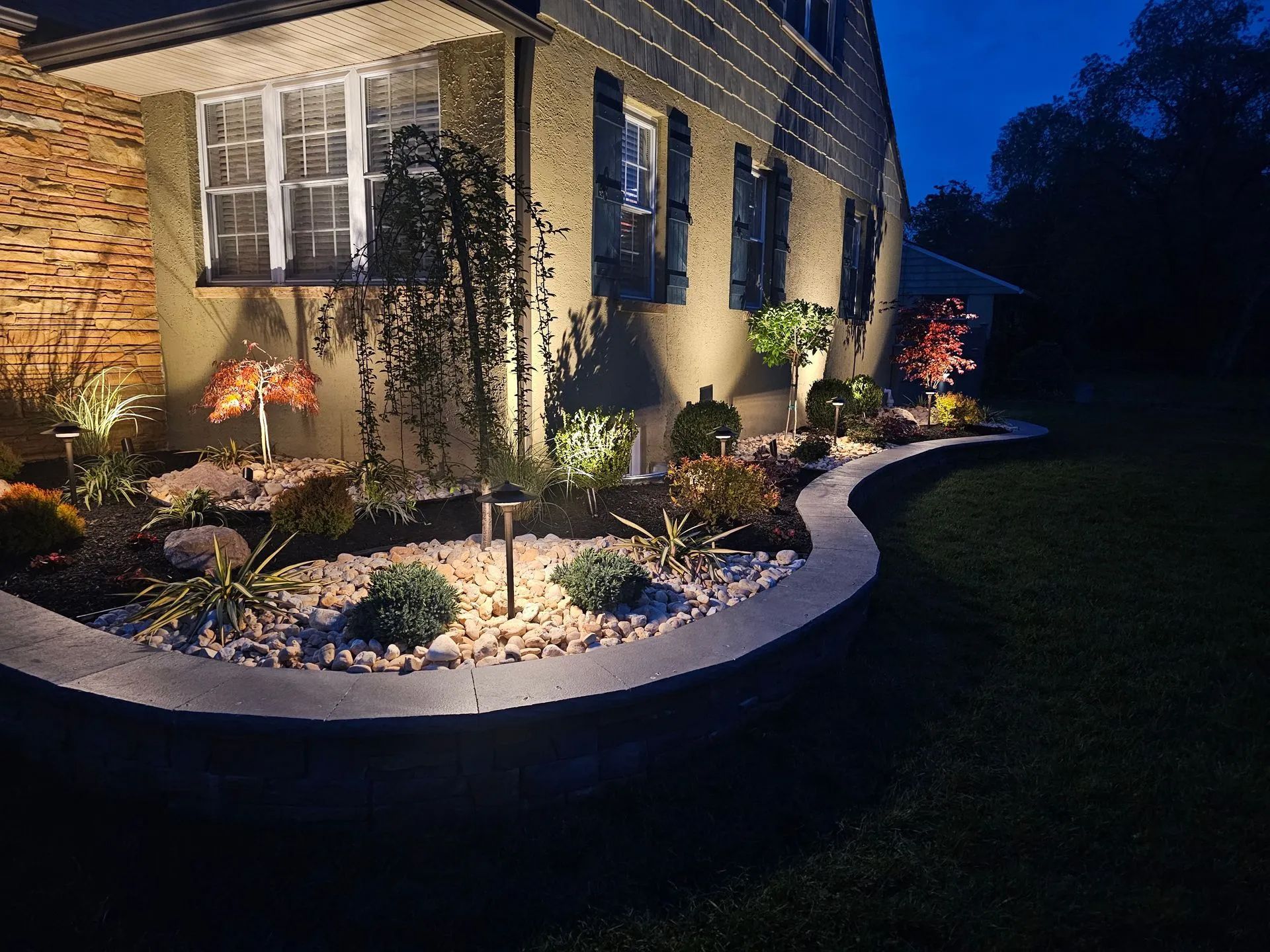 A curved stone garden bed with illuminated shrubs and trees sits in front of a house at twilight.