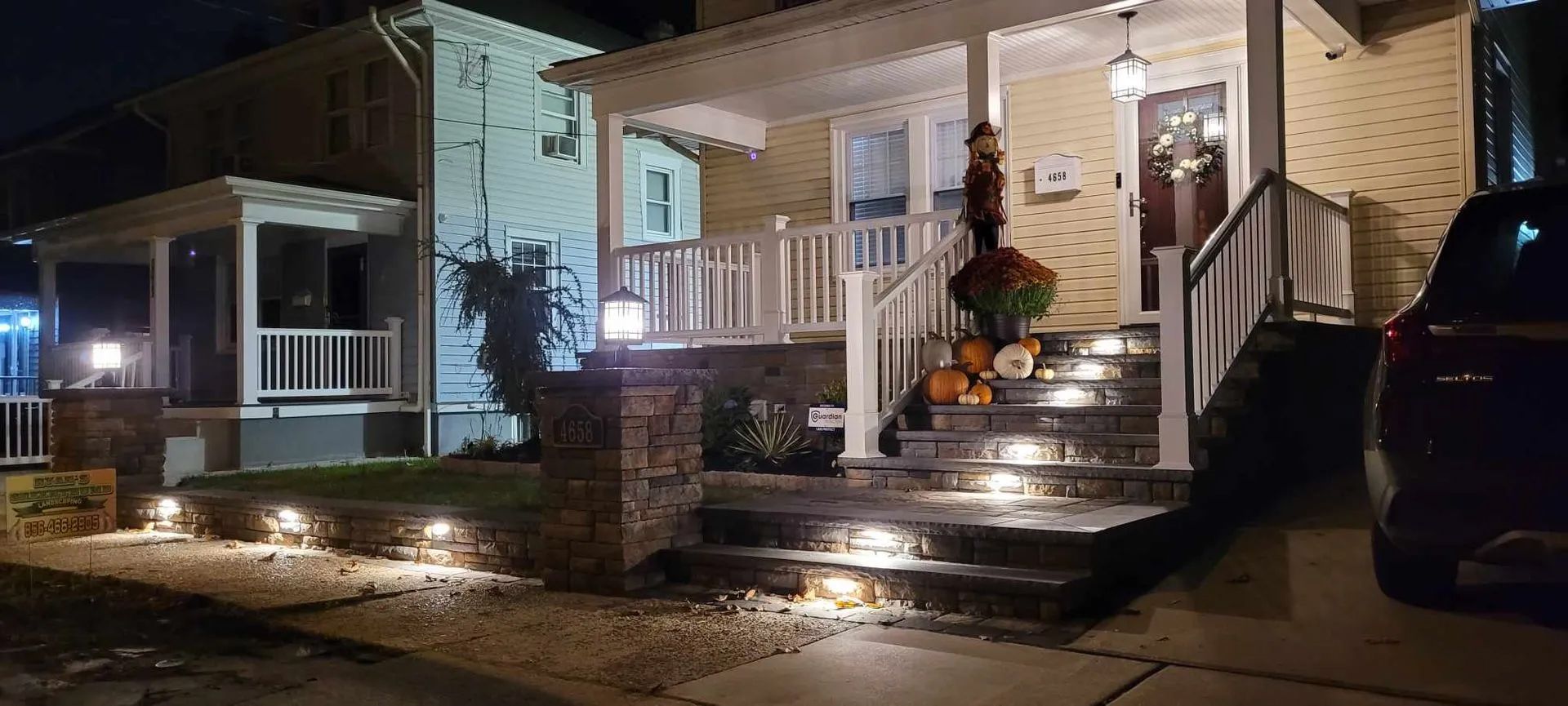 A yellow house at night with a stone retaining wall and illuminated porch steps, accented by pumpkins near the entrance.