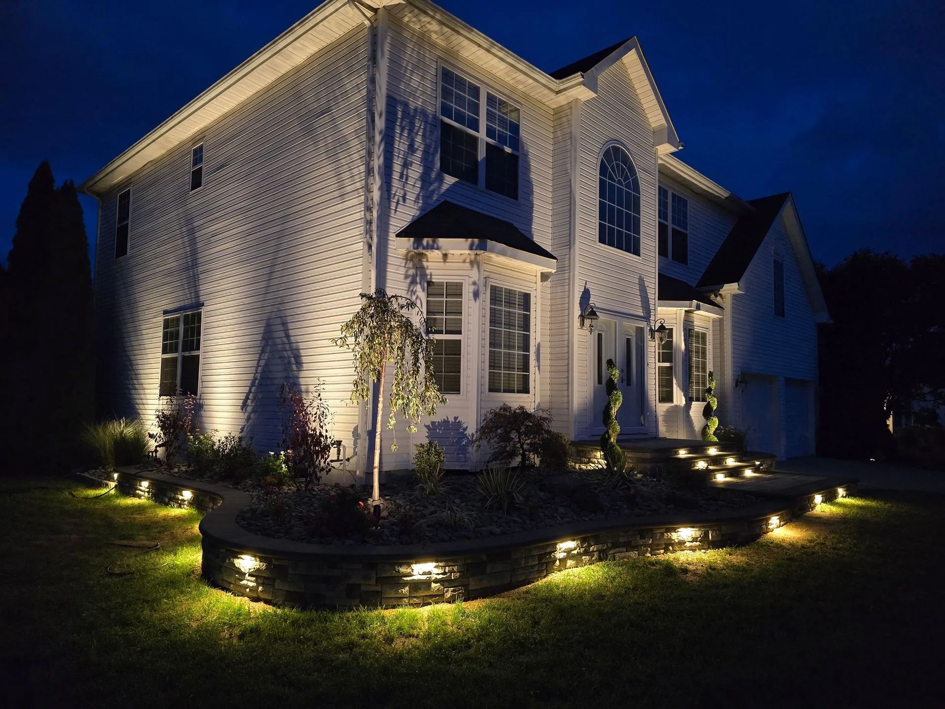 A two-story white house at night, illuminated by warm landscape lighting along the front garden and stone retaining wall.