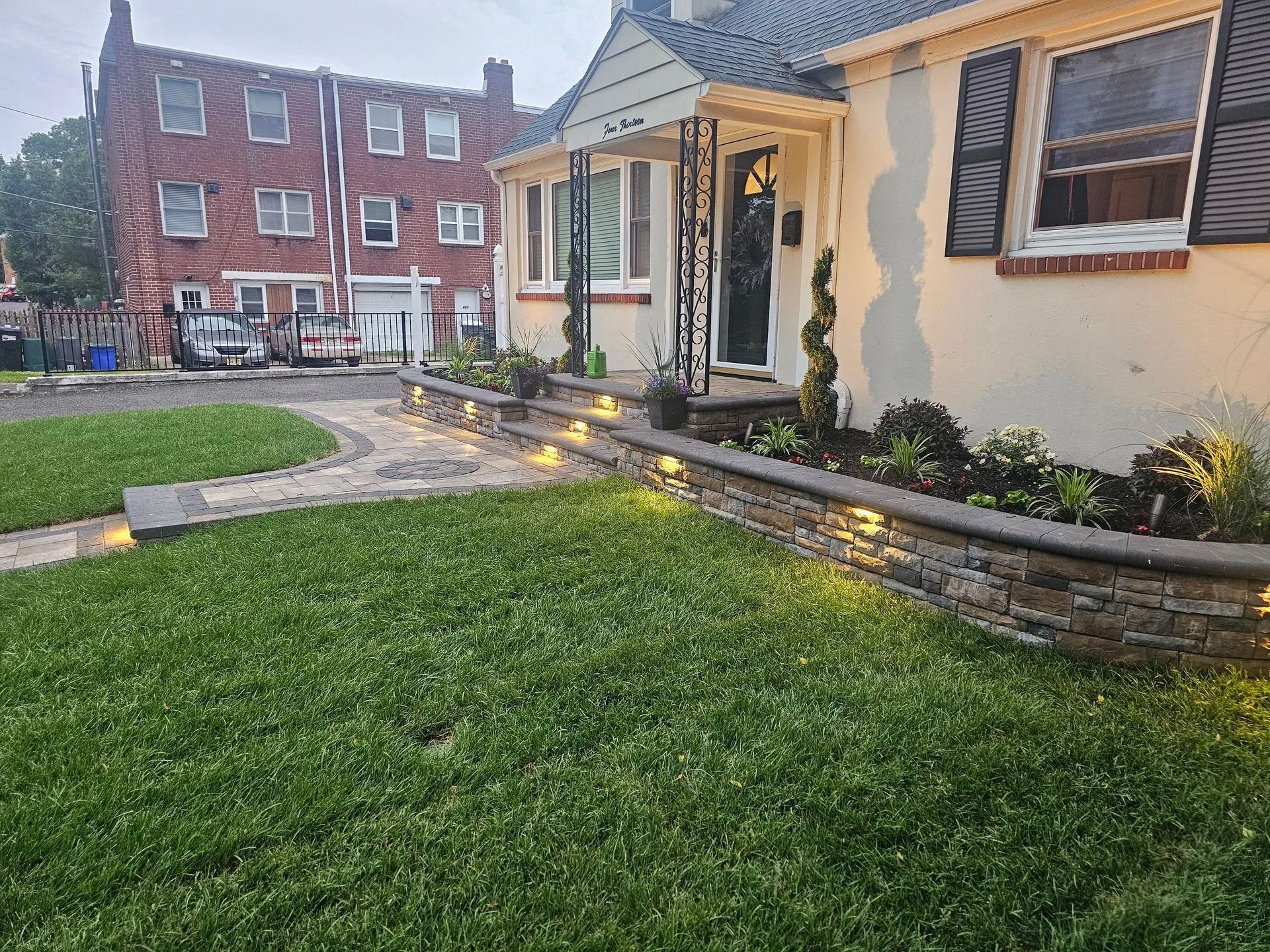 A suburban house at dusk with a tiered stone retaining wall and walkway lit by small embedded warm lights.