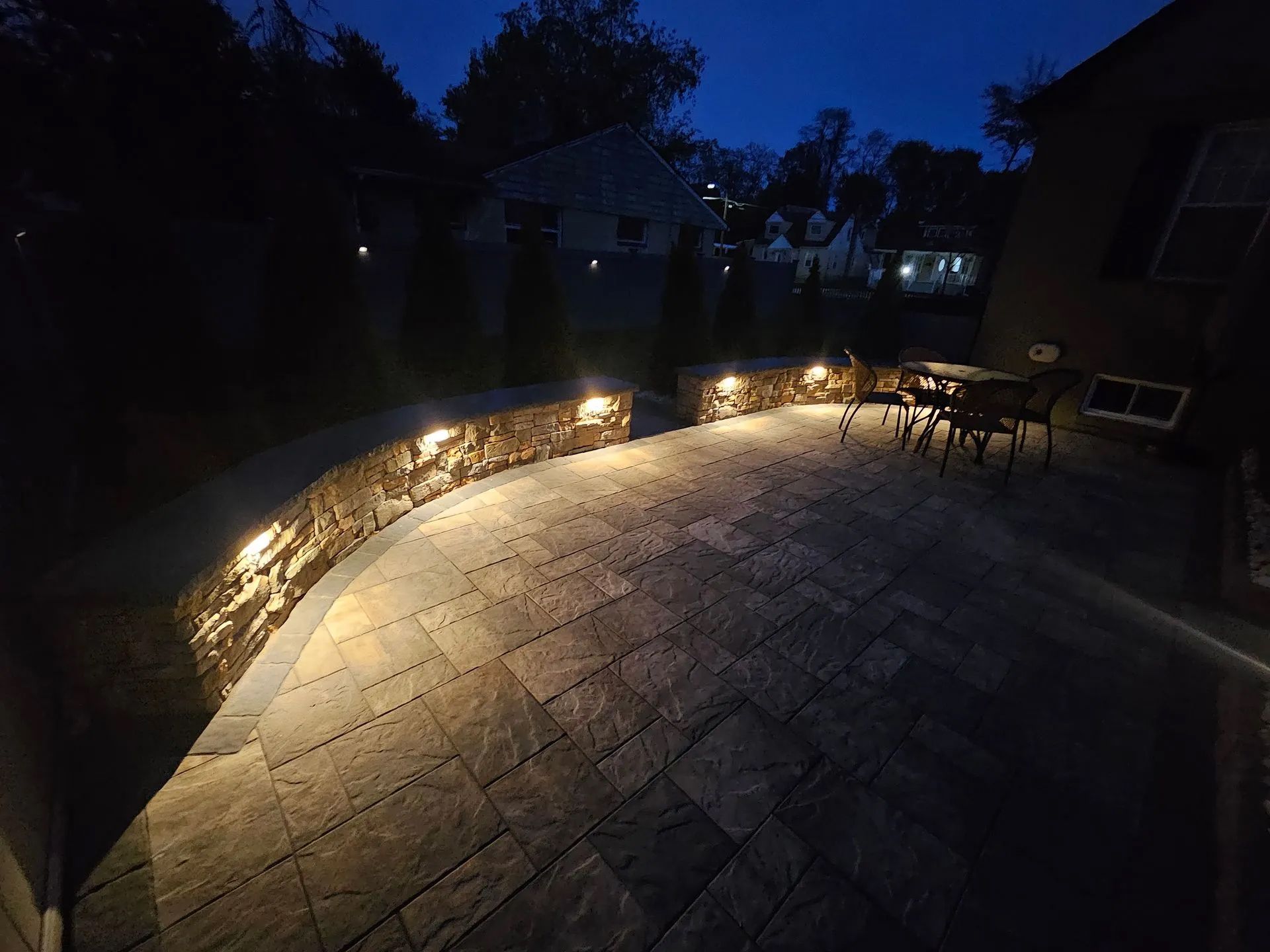 A stone patio at dusk, illuminated by warm recessed lights set into a curved retaining wall.