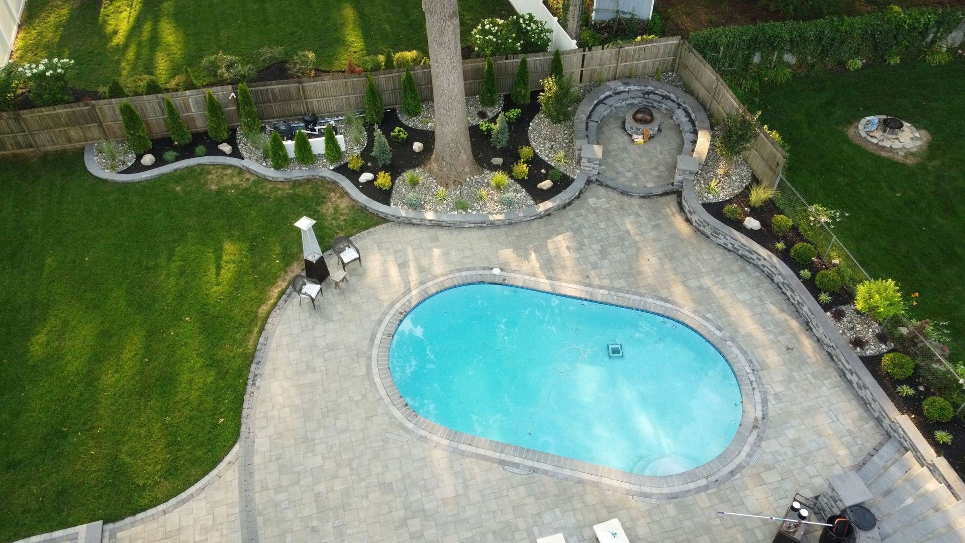 An aerial view of a backyard featuring a turquoise oval pool, stone patio, and a fire pit area surrounded by landscaping.