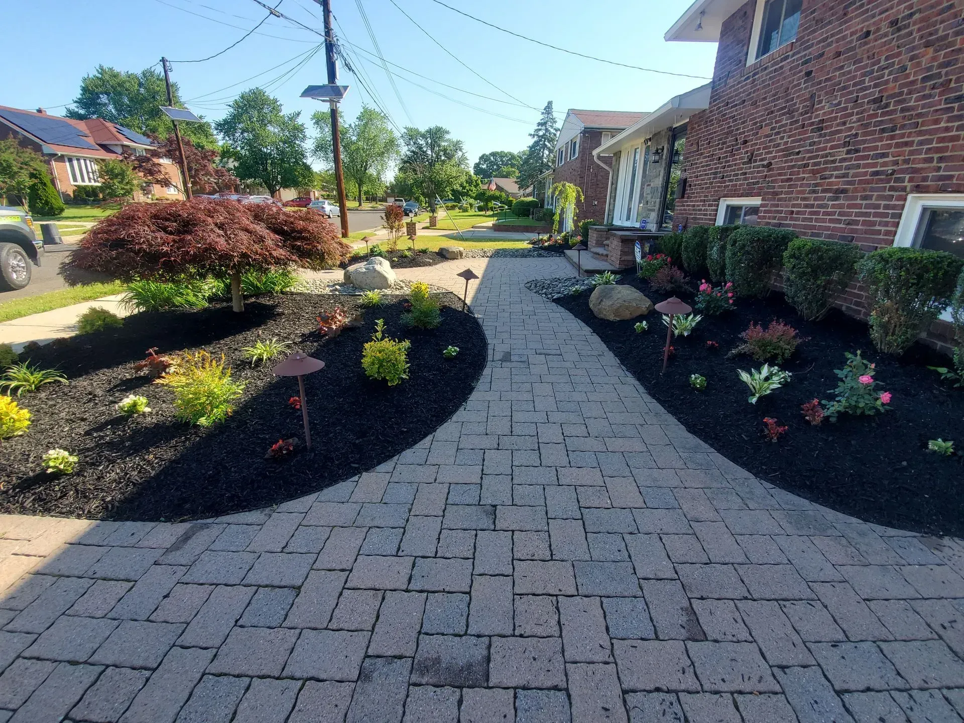 A paved walkway leads to a brick house, flanked by landscaped garden beds with black mulch, shrubs, and small plants.