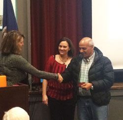 Three people shake hands at a presentation, with a woman holding a certificate beside a podium.