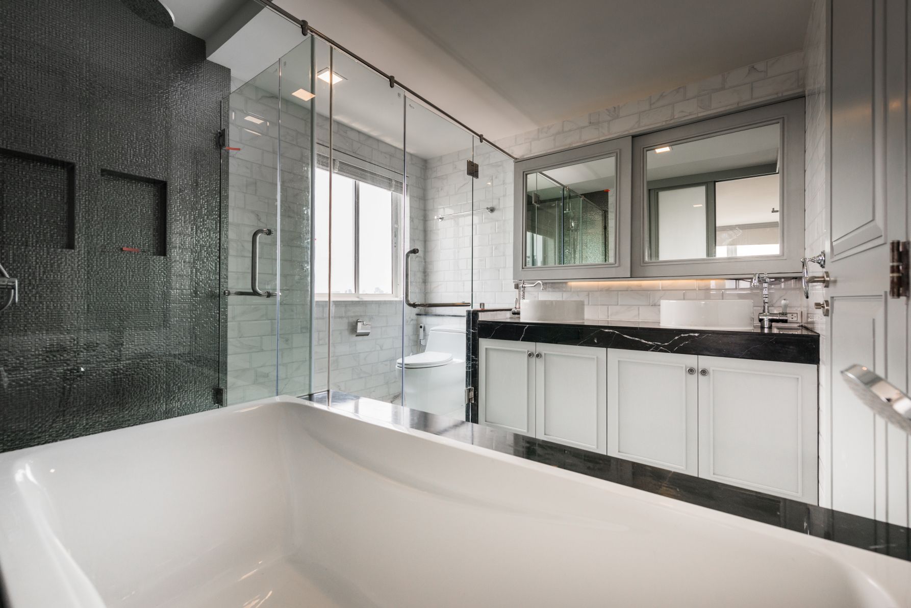 A modern, bright bathroom with a white freestanding tub, black marble vanity, double mirrors, and a glass-enclosed shower.