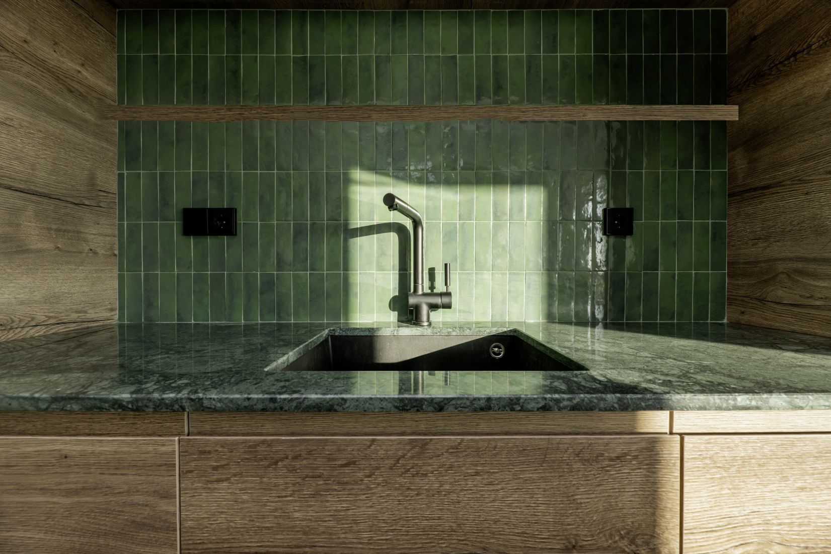 A modern kitchen sink with a dark faucet, set against green rectangular tiles and wood cabinets under sunlight.