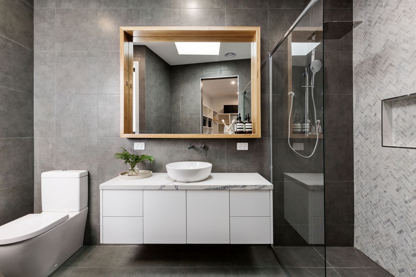 A modern bathroom with grey tiled walls, a white floating vanity, a round vessel sink, and a wooden-framed mirror.
