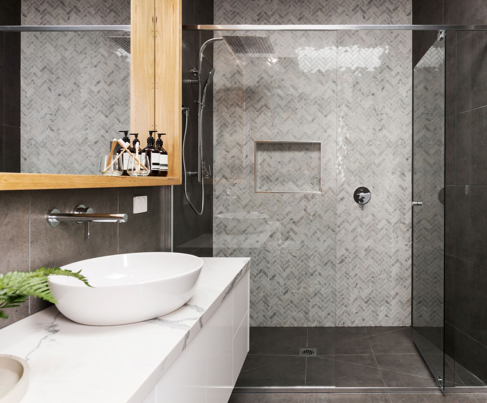 Modern bathroom with a white vessel sink on a stone vanity, beside a walk-in shower with herringbone tiles and glass door.