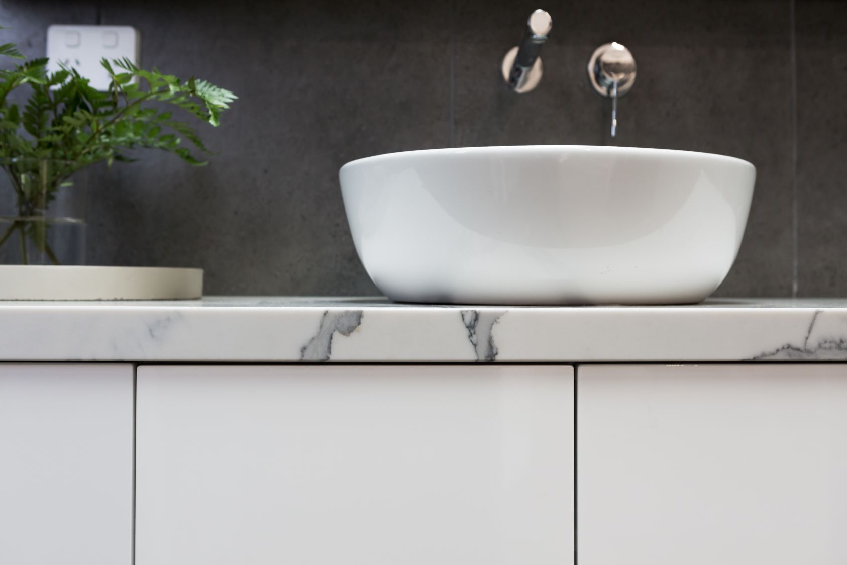 A white vessel sink sits on a marble countertop in front of a dark tiled wall, with a small potted plant to the left.